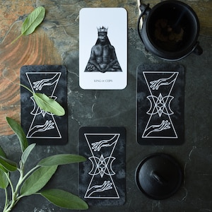 May include: Three tarot cards on a dark gray background. The top card is white with a black and white illustration of a king with a crown and a cup. The text on the card reads "King of Cups". The two cards below are black with white illustrations of hands and a geometric design.