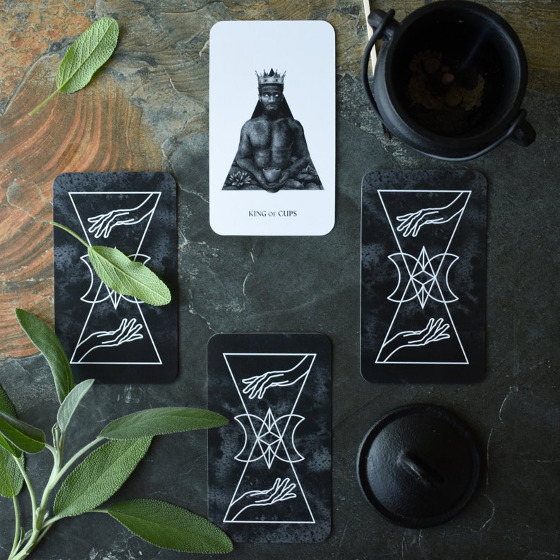 May include: Three tarot cards on a dark gray background. The top card is white with a black and white illustration of a king with a crown and a cup. The text on the card reads "King of Cups". The two cards below are black with white illustrations of hands and a geometric design.