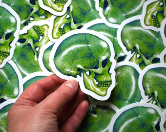 May include: A hand holding a green skull sticker with a white border. The sticker has a detailed illustration of a skull with shading in shades of green and blue. Other similar stickers are in the background.