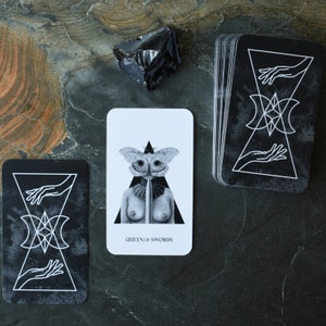 May include: A black and white tarot card with a woman with a moth head and a sword. The card reads "Queen of Swords".