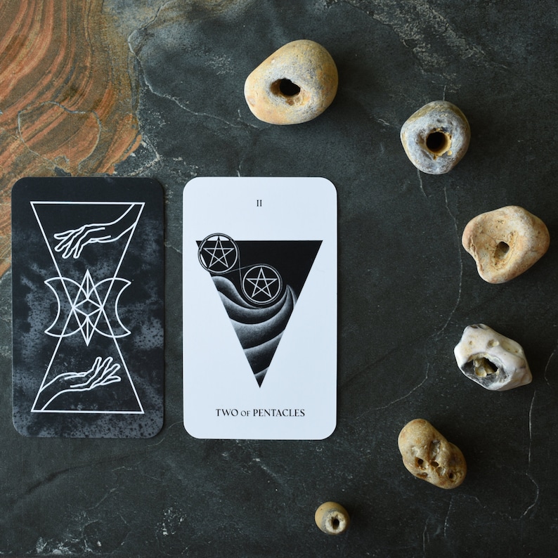 May include: Two of Pentacles tarot card with black and white illustrations of hands, a pentagram, and a triangle. The card is on a dark gray surface with several small, light brown stones.