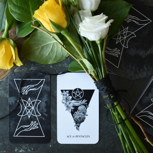 May include: Two tarot cards, one with a black and white illustration of a pentagram and the other with a black and white illustration of a hand with a pentagram. The cards are on a black surface with a bouquet of white and yellow roses.