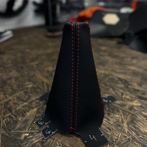 May include: Black leather gear shift boot with red stitching. The boot is cone-shaped and has a flat base.