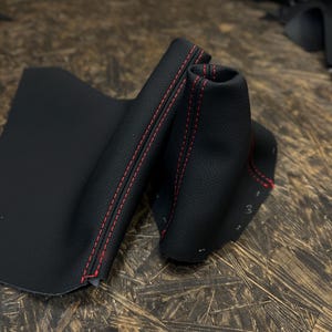 Puede incluir: Funda de palanca de cambios de cuero negro con costuras rojas. La funda está doblada por la mitad y las costuras son visibles en ambos lados.