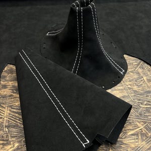 Könnte beinhalten: Zwei schwarze Schaltknauf-Abdeckungen aus Wildleder mit weißen Nähten. Die Abdeckungen sind aus weichem, strapazierfähigem Wildleder gefertigt und haben ein klassisches Aussehen.