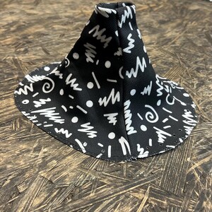 Peut inclure: Un soufflet de levier de vitesses en tissu noir avec un motif abstrait blanc de lignes, de points et de spirales. Le soufflet de levier de vitesses est en forme de cône et possède une base circulaire avec de petits trous sur le bord.