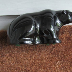 May include: A black ceramic bear figurine with a glossy finish. The bear is sitting on its haunches with its head turned to the side. The figurine is on a brown surface.