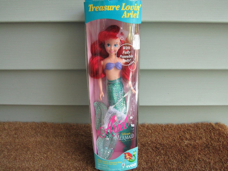 May include: A plastic doll of Ariel from Disney's The Little Mermaid in a green mermaid tail with a purple seashell bra. The doll is packaged in a clear plastic box with a blue and white background. The box says "Treasure Lovin' Ariel" and "With Fully Poseable Body!"