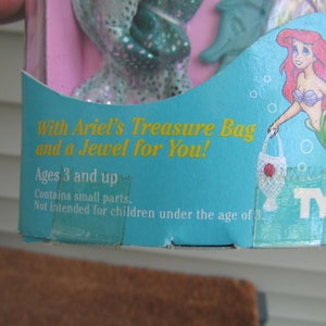 May include: A blue and green plastic toy box with a picture of Ariel from The Little Mermaid. The box says "With Ariel's Treasure Bag and a Jewel for You!" and "Ages 3 and up".