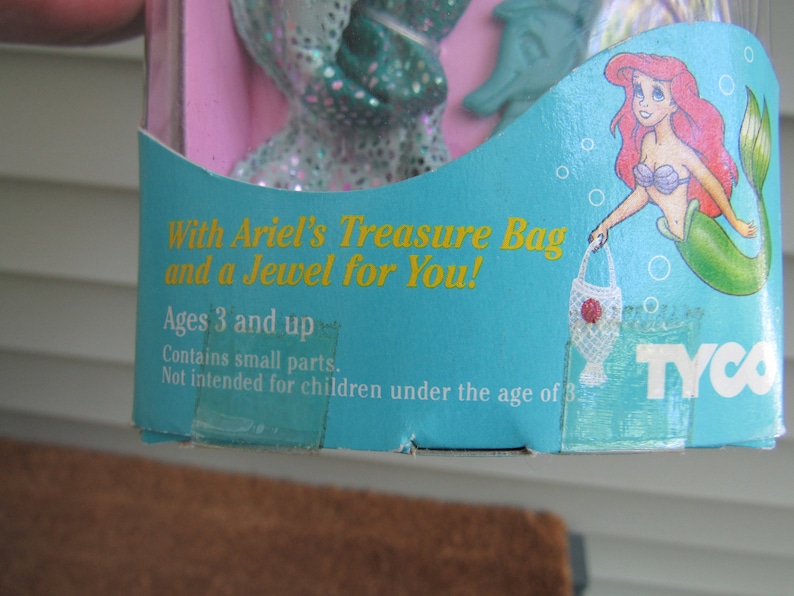 May include: A blue and green plastic toy box with a picture of Ariel from The Little Mermaid. The box says "With Ariel's Treasure Bag and a Jewel for You!" and "Ages 3 and up".