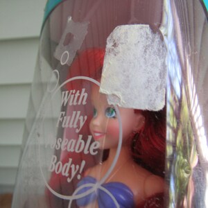 May include: A red-haired doll with a purple top and a white seashell necklace. The doll is in a clear plastic package with the text "With Fully Poseable Body!" printed on it.
