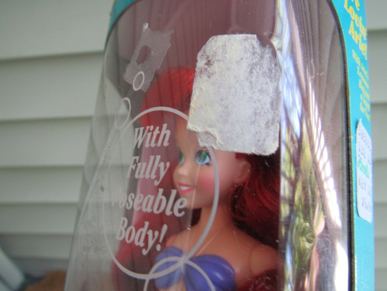 May include: A red-haired doll with a purple top and a white seashell necklace. The doll is in a clear plastic package with the text "With Fully Poseable Body!" printed on it.