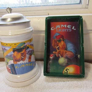 May include: A white ceramic beer stein with a silver lid and a colorful illustration of a camel playing an accordion. Beside it is a green ashtray with a Camel Lights advertisement featuring a camel playing pool.
