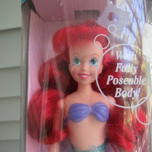 May include: A red-haired Ariel doll with a purple bikini top and a green tail. The doll is in a plastic package with the text "With Fully Poseable Body!" on it.