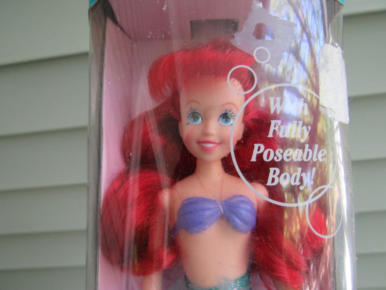 May include: A red-haired Ariel doll with a purple bikini top and a green tail. The doll is in a plastic package with the text "With Fully Poseable Body!" on it.
