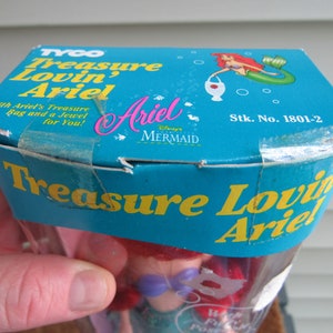 May include: A blue and yellow box with the words "Treasure Lovin' Ariel" on it. The box contains a Disney's The Little Mermaid Ariel doll. The box also has the words "Stk. No. 1801-2" on it.