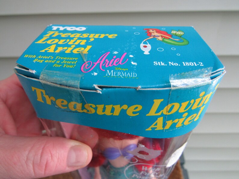 May include: A blue and yellow box with the words "Treasure Lovin' Ariel" on it. The box contains a Disney's The Little Mermaid Ariel doll. The box also has the words "Stk. No. 1801-2" on it.