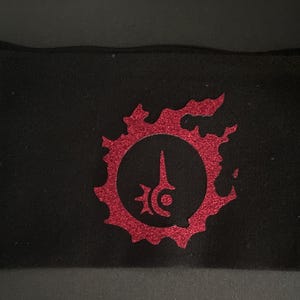 May include: Black fabric pouch with a red, glittery emblem. The emblem is a circular design with a central vertical element and surrounding stylized shapes. The pouch is rectangular and appears to be made of a soft material.