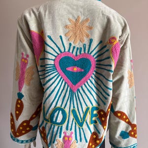 May include: A light gray jacket featuring colorful embroidery on the back. The design includes a central blue heart with a pink border, a sunburst, and the word LOVE in blue letters. Additional motifs include pink butterflies, orange carrots, and brown shapes.