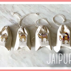 May include: Four small, beige drawstring bags with embroidered camels. Each camel is decorated with colorful saddle blankets and tassels. The bags are arranged in a row on a white marble surface. The word "JAIPUR" is printed in white letters on a pink heart-shaped background below the bags.