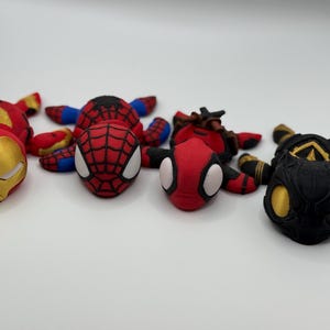 May include: A collection of superhero-themed figurines, including Iron Man, Spider-Man, and Deadpool. The figurines are primarily red, black, and gold, with detailed facial features and costume designs. They are arranged on a white surface.