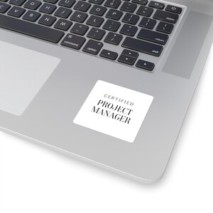 May include: A white square sticker with the text "CERTIFIED PROJECT MANAGER" in black, placed on a silver laptop. The laptop keyboard is visible, with black keys and a silver frame.