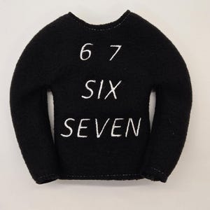May include: Black sweater with white embroidered text. The text reads "6 7 SIX SEVEN". The sweater has long sleeves and a rounded neckline. The background is plain white.