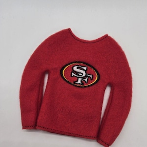 May include: A red long-sleeved shirt with a crew neck and a team logo patch. The patch is an oval shape with a gold border, and features the letters "SF" in white, with a red background.
