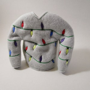 May include: A gray felt sweater-shaped ornament decorated with embroidered colorful Christmas lights. The lights are red, blue, yellow, and purple, strung along green lines. The ornament has a V-neck and two sleeves.