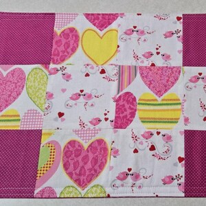 May include: A pink and white patchwork table runner with a heart and bird design. The runner is made of different fabrics with various patterns, including polka dots, stripes, and floral prints.