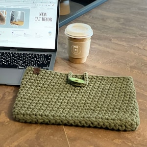 May include: A green crocheted laptop sleeve with a wooden button closure. The sleeve is sitting on a wooden table next to a laptop computer and a cup of coffee. The laptop screen shows an Etsy shop page for cat decor.