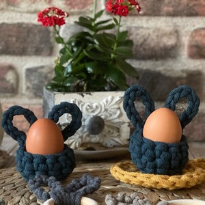 May include: Two crocheted egg holders shaped like bunnies with brown eggs inside. The holders are teal blue and are sitting on a woven placemat with a green crocheted coaster and a wooden ring.