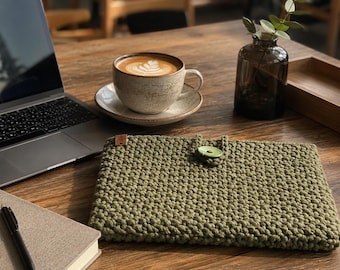 May include: A rectangular olive green crochet laptop sleeve featuring a textured stitch pattern, a small brown leather tag on the top left corner, and a single light green button closure centered on the top edge for secure storage.