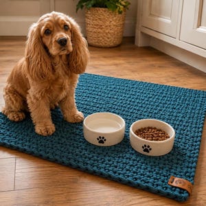 Pode incluir: Um tapete retangular de malha grossa em azul-petróleo escuro com uma pequena etiqueta de couro castanho no canto. Duas taças redondas de cerâmica esbranquiçada com estampas de patas pretas estão sobre o tapete, uma contém ração seca castanha.