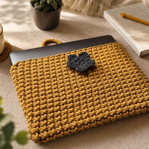 Puede incluir: Una funda de ganchillo para portátil en color amarillo mostaza con un patrón de puntos texturizados y un adorno de flor de ganchillo gris oscuro centrado en la solapa frontal. La funda es rectangular y diseñada para un dispositivo fino.