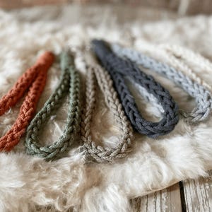 May include: Braided rope necklaces in various colors, including orange, green, gray, and blue, are displayed on a fluffy, white surface. The necklaces are made of thick, woven rope, showcasing a textured design. The neutral background enhances the colors of the necklaces.