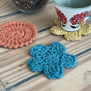 May include: Three crocheted coasters in different colors and shapes. A round orange coaster, a flower-shaped blue coaster, and a flower-shaped yellow coaster.