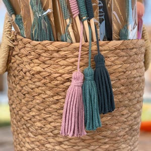 May include: A woven straw basket filled with multiple packaged wands, each featuring a long wooden handle wrapped in colored cord and finished with a hanging tassel in shades of dusty pink, teal, and dark navy blue, displayed in a vertical arrangement.