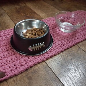 May include: A pink crocheted pet mat with a black metal bowl filled with dry pet food and a clear glass bowl for water.