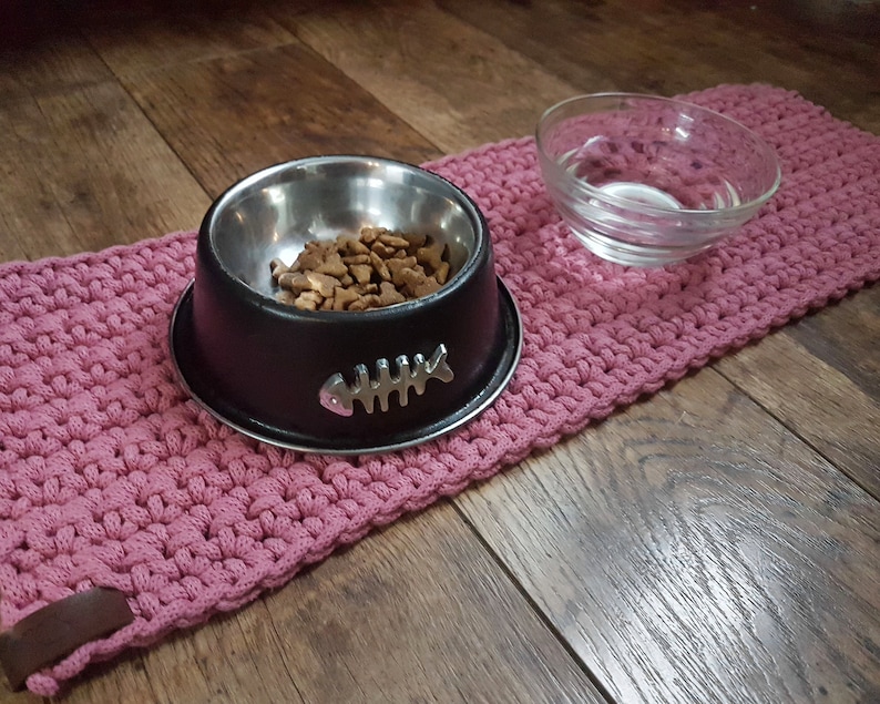 May include: A pink crocheted pet mat with a black metal bowl filled with dry pet food and a clear glass bowl for water.