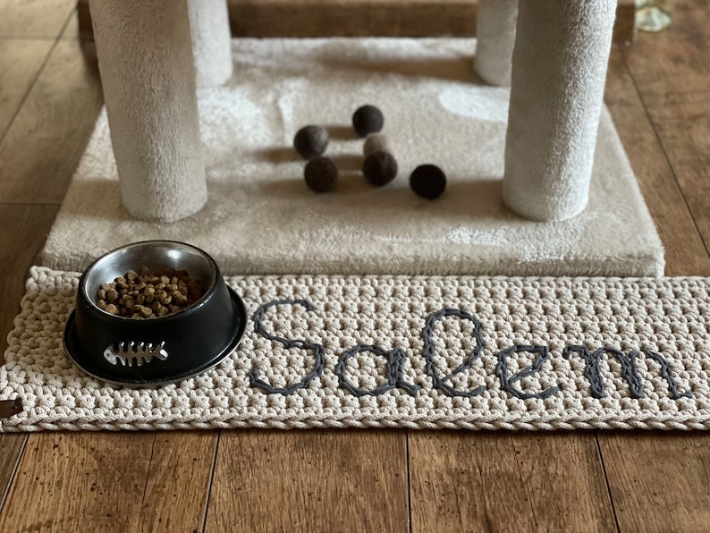 May include: A beige crocheted pet mat with the name "Salem" embroidered in grey. A black metal bowl with a fishbone design is filled with brown pet food.