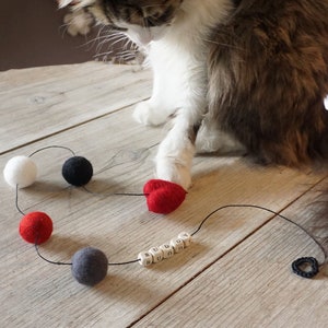 Könnte beinhalten: Eine Katze spielt mit einem roten herzförmigen Filzspielzeug an einer Schnur. Die Schnur hat auch schwarze, weiße und graue Filzkugeln und Holzkugeln, die "BUDDY" buchstabieren.