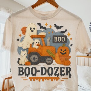 Op de afbeelding: Crèmekleurig T-shirt met een Halloween-thema. Het ontwerp bevat een cartoon tractor met geesten, een pompoen en de tekst "BOO-DOZER". De afbeelding is in oranje, blauw en zwart.