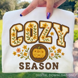 Könnte beinhalten: Weißes Sweatshirt mit dem gestickten Schriftzug "COZY SEASON" in Braun und Gelb. Das Wort "COZY" ist mit gelben Blumen und grünen Blättern verziert. In der Mitte befindet sich ein Kürbis. Die Worte "DIGITAL DOWNLOAD ONLY" stehen unten.