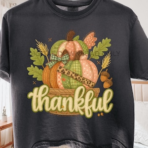 Puede incluir: Camiseta gris oscuro con un diseño temático de otoño. El diseño incluye calabazas en tonos naranja, verde y rosa, con un estampado de leopardo. La palabra "thankful" está escrita en una fuente decorativa.