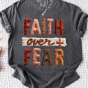 May include: Dark grey t-shirt featuring the embroidered words "FAITH over FEAR". The text is in shades of brown, orange, and cream. A silver necklace with a pendant is visible.