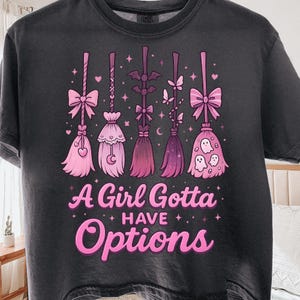May include: Dark gray t-shirt featuring five pink witch brooms with bows, hearts, and ghost designs. The text "A Girl Gotta Have Options" is printed in pink and white. A fun and whimsical design for Halloween or everyday wear.