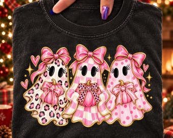 May include: A charcoal gray t-shirt featuring a graphic of three ghosts wearing large pink bows. Each ghost holds a small pink pumpkin and displays a different pattern: leopard print, pink and white checkered, and pink polka dots.