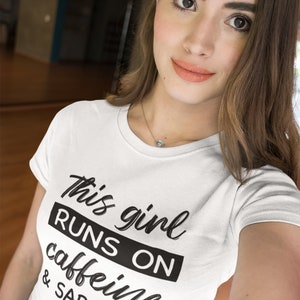 May include: A white short-sleeved t-shirt featuring the black printed text "This girl RUNS ON caffeine & SARCASM". The design uses a mix of cursive and bold block lettering, with the word "RUNS ON" highlighted inside a solid black rectangular box.