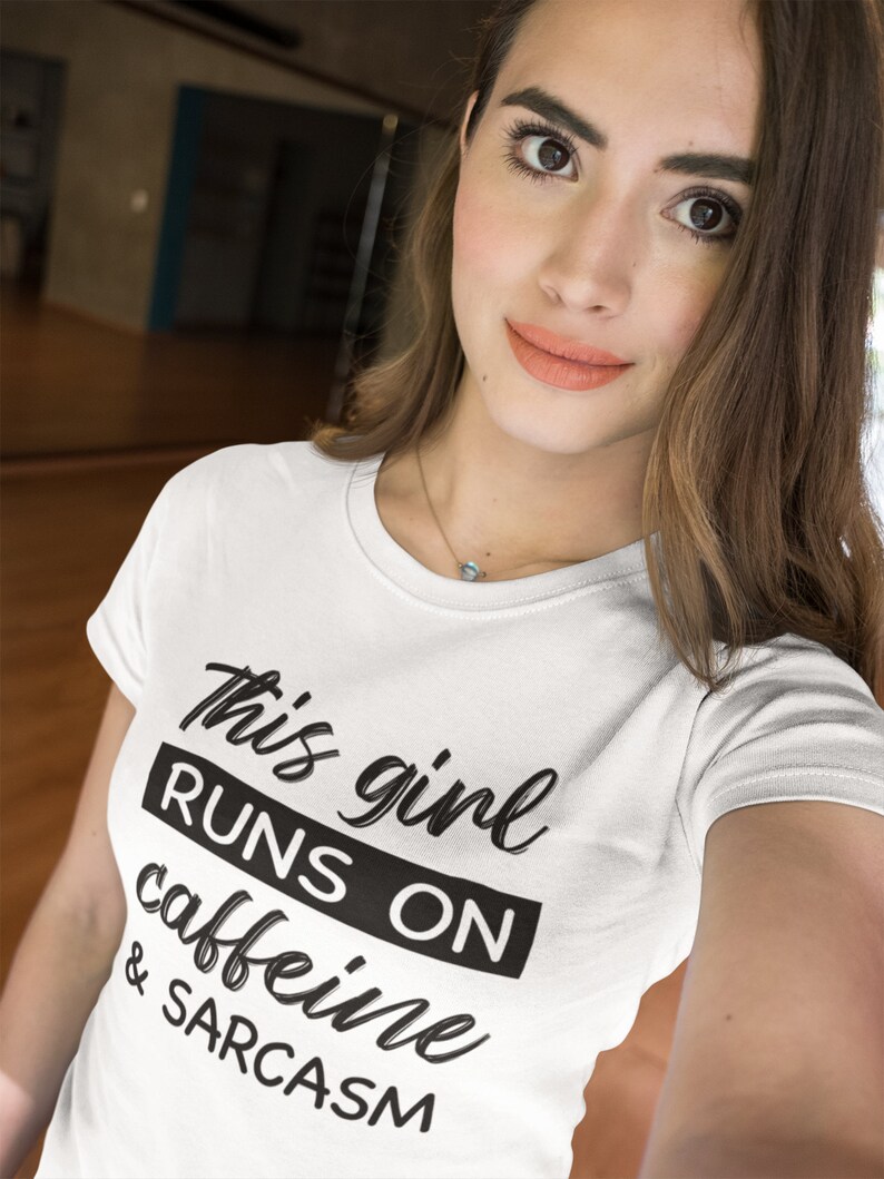 May include: A white short-sleeved t-shirt featuring the black printed text "This girl RUNS ON caffeine & SARCASM". The design uses a mix of cursive and bold block lettering, with the word "RUNS ON" highlighted inside a solid black rectangular box.